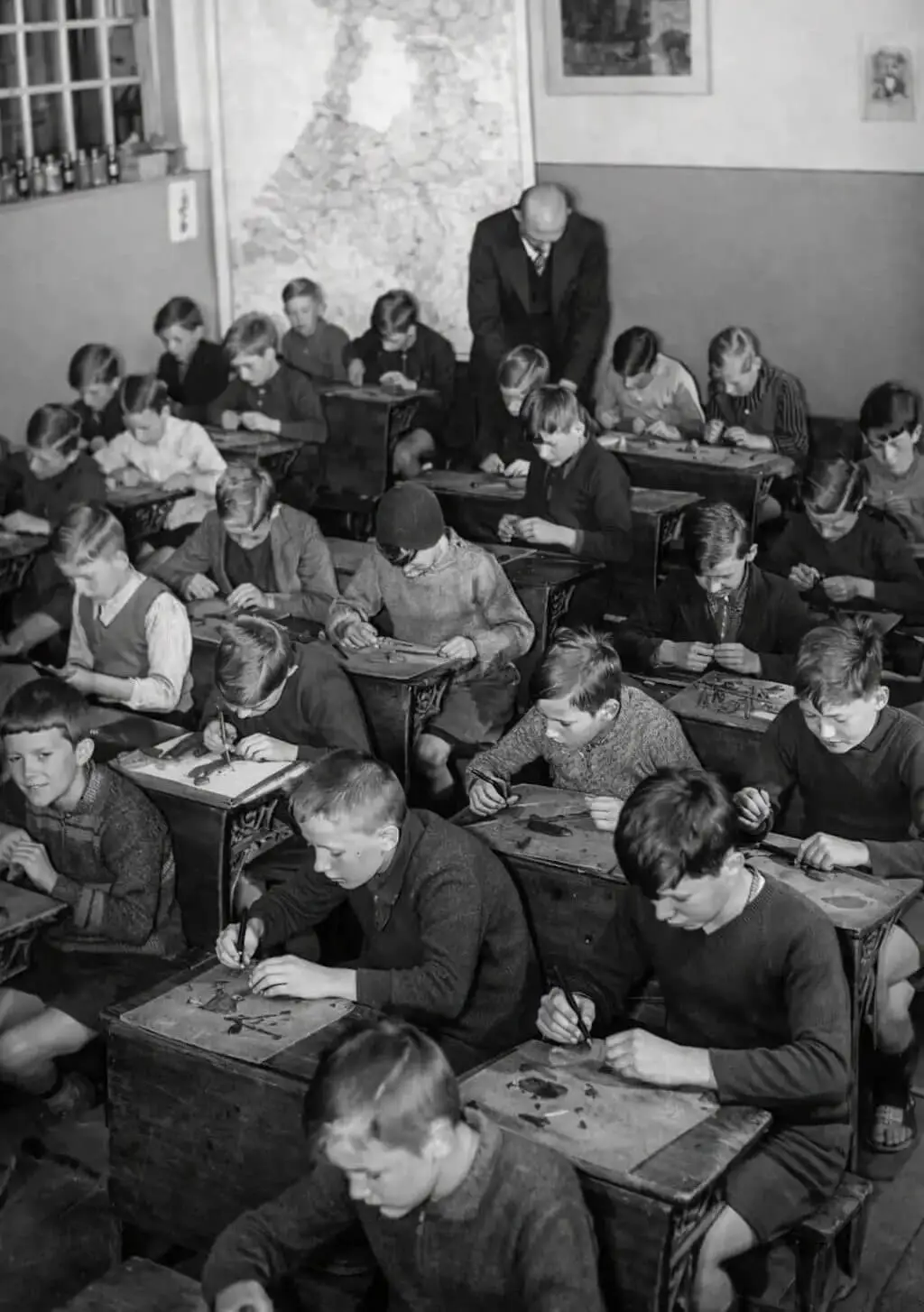 Kinderen zitten in een ouderwets klaslokaal aan houten schoolbanken terwijl ze handvaardigheidsoefeningen doen, met een leraar die achterin toezicht houdt.